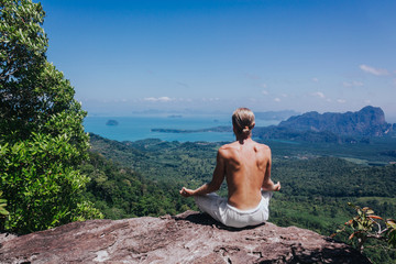 Back view of shirtless male practicing yoga. Back view of young man sitting on rock cliff in lotus pose on background of tropical valley.