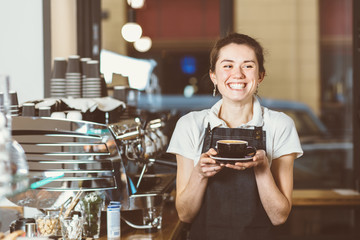 young female barista adding milk to coffee and smiling while standing at the bar counter in the urban cafe - obrazy, fototapety, plakaty