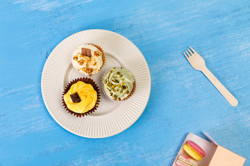 Cupcakes with buttercream. Macaroons in box. Sweet cake with pistachio nuts cream and small chocolate bar. Dessert with topping. Wooden background.