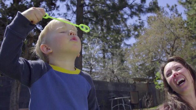Adorable little boy gets a big breath and tries blowing bubbles using a wand in his backyard, eventually he succeeds, his mom laughs with joy