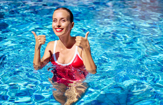 Happy summer vacation. Smiling young woman in red swimsuit showing thumbs up at swimming pool.