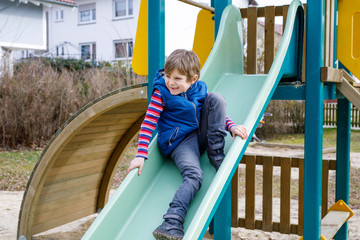 Happy blond kid boy having fun and sliding on outdoor playground