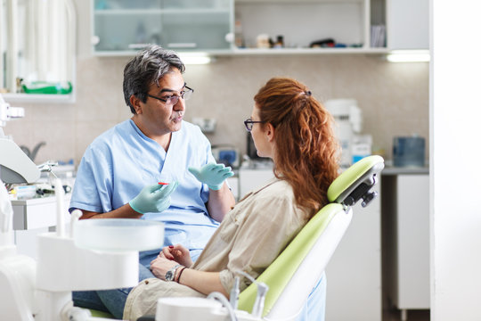 A professional dentist, equipped with a bright smile, converses with his red-haired female patient, carefully explaining the upcoming treatment, and ensuring her comfort throughout the process.