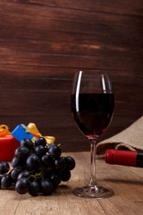 Red wine bottle. Wineglass with dark grapes branch. Gift box with bow on rustic wooden background.