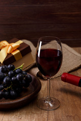 Red wine bottle. Wineglass with dark grapes branch. Gift boxes with bows on rustic wooden background.