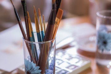 Paint brushes in glass on the table in a workshop. Master drawing at background.Close up.