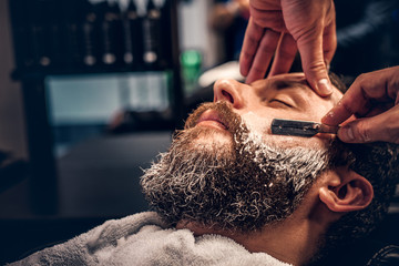 Close up image of barber shaving a man with a sharp steel razor. - obrazy, fototapety, plakaty