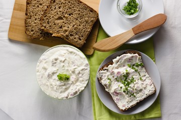 Slice of wholegrain rye bread with delicate light and fresh cottage cheese spread sprinkled with chives and parsley. Wooden spreading knife and some extra herbs in glass cup