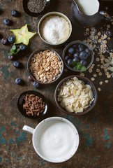 Obraz na płótnie Canvas Ingredients for making smoothie for healthy breakfast. Bowls of yogurt, blueberries, granola, almond chia seeds, coconut, milk, chocolate, mint, carambola over dark wooden background. Top view, space