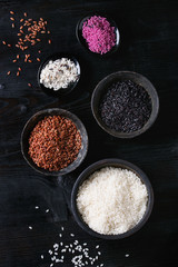 Variety assortment of raw uncooked colorful rice white, black, brown, pink in black bowls over burnt wooden background. Top view with space