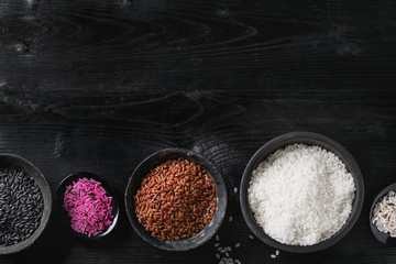 Obraz na płótnie Canvas Variety assortment of raw uncooked colorful rice white, black, brown, pink in black bowls over burnt wooden background. Top view with copy space