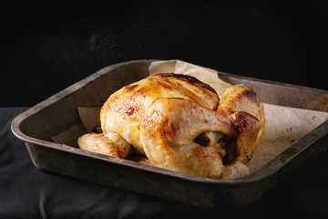 Grilled baked whole organic chicken on backing paper in old oven tray over black burnt wooden background. Top view with space.