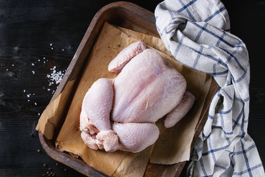 Raw Organic Uncooked Whole Chicken With Salt And Pepper On Backing Paper In Old Oven Tray With White Kitchen Towel Over Black Burnt Wooden Background. Top View With Space.