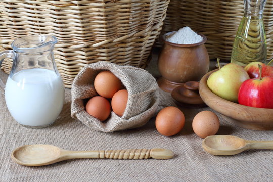Composition of products for baking and eating. Milk, flour, oil, eggs, apples are on the table in the background of wicker baskets.