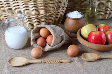 Composition of products for baking and eating. Milk, flour, oil, eggs, apples are on the table in the background of wicker baskets.
