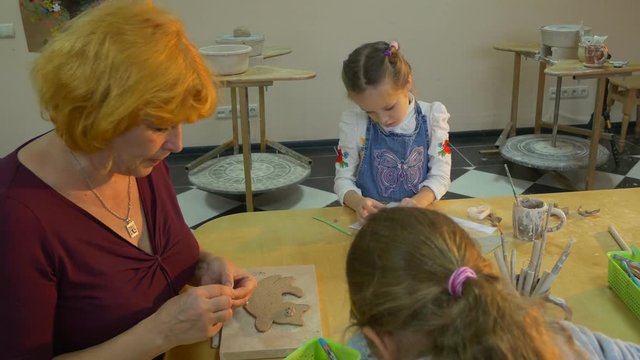 Two Girls and a Professional Master Are Making Clay Toys While Sitting at a Big Square Table, Covered With a Lot of Drawings, Boards and Thin Sticks