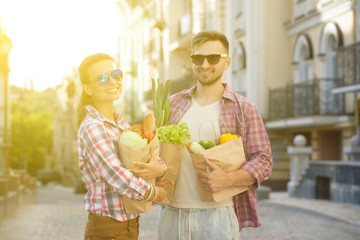 Fototapeta premium Happy couple after shopping. Man and woman in sunglasses carrying bags full of different food. Toned image.