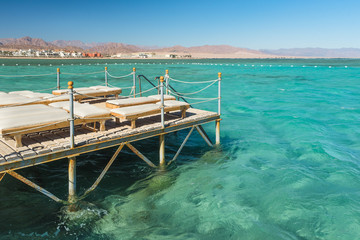 Pier with chaise longues in the sea in resort. Summer vacation. View at a clear sea with turquoise water. Summer vacation at a sea coastline in an exotic country. Staircase to the water for swimming.