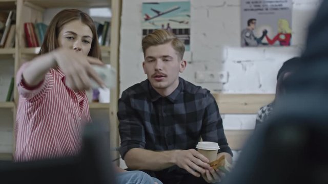 Tracking shot of young smiling man and woman taking selfie with smart phone at break in the office. Man holding coffee cup and sandwich; their colleague working on laptop in the background