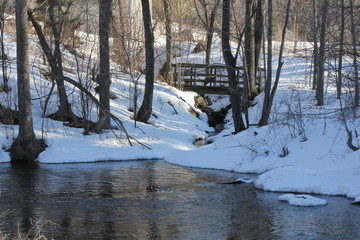 Little wooden bridge spanning a deep crevice full of rocks, water, and snow, in an area filled with snow and trees.  
