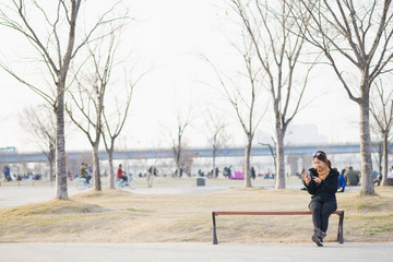 Fototapeta na wymiar Cute asian woman chatting, reading and checking text massage on smartphone while sitting on wood bench in the park. Feeling happy and chilling.