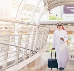 Arab man in travel concept . Young saudi arabic man in traditional clothes walking with suitcase on...