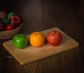 Green apple, red apple and orange on chopping block and fruits in a basket on desk background