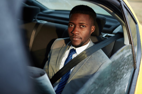 Portrait of confident African -American businessman riding in backseat of car  looking at camera out of window lit by sunlight, while reading newspaper