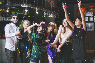 Young cheerful company of friends in the club bar having fun with multi-colored confetti and...