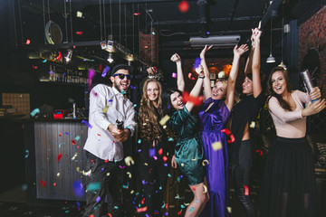 Young cheerful company of friends in the club bar having fun with multi-colored confetti and...