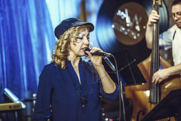 A European middle-aged female singer in a cap sings into the microphone on the restaurant scene.