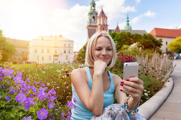 Female traveler in a blue T-shirt and a long skirt sitting and looking in a mobile phone on the background of Wawel Castle. Krakow, Poland