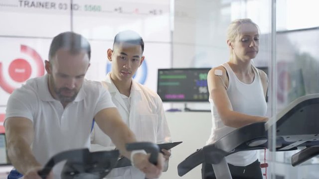  Man & woman using fitness machines being tested and monitored by sports scientist. 