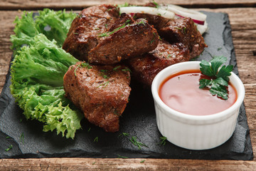 Delicious shashlik served with lettuce salad and onion rings on black slate with tomato sauce, on rustic wooden table, close up view. Restaurant menu photo.