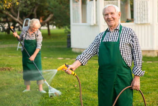 Man with garden hose smiling. Senior male in apron outdoors. Find jobs in gardening.