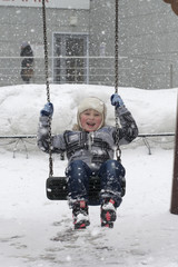 The happy little boy shakes on a swing in the winter in snowfall.