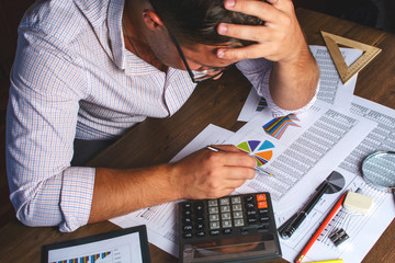 The man (businessman, manager, analyst, student) working with tables, graphs, diagrams, makes calculations at the wooden table in the office in the evening (or at night), after hours