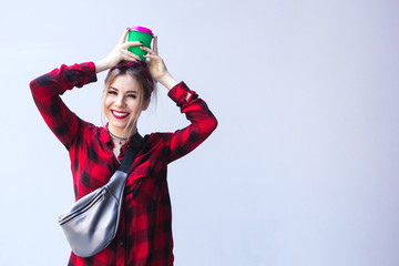 Fototapeta premium A blond hipster girl in a red shirt in a square and a bandanna stands near a wall, smiling and holding a glass with coffee on her head.