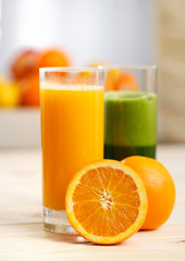 Orange juice and green vegetable juice in tall glasses with an orange half and a basket of fruit in the background
