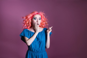 girl with pink hair in blue dress shows sign on lilac background, place for text
