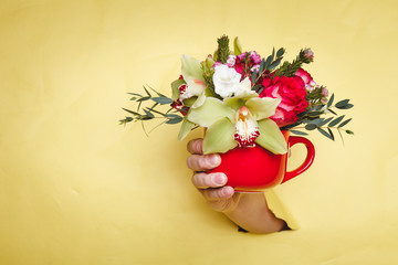 Hand holds small vase with flowers and lily breakes through a yellow background