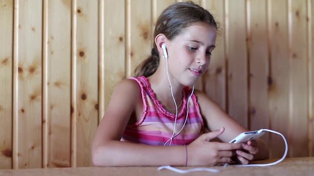 Girl with smartphone sits at table, listens to music and sings a song. Girl with white smartphone and earpieces listens to music, gesticulates and sings a song