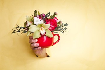 Hand holds small vase with flowers and lily breakes through a yellow background