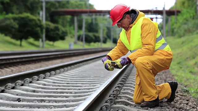 Railway worker in yellow uniform with level measuring instrument on railway line. Railway man in red hard hat sits on railway tracks and looks at the train. Workman with level measuring instrument