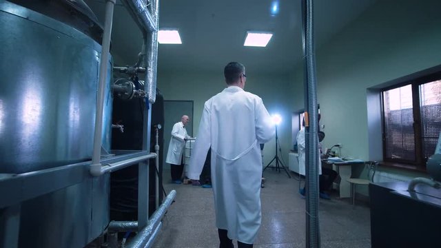 Rear wide view video of unrecognizable male scientist in white uniform walking through lad with metal tubes and engineering equipment aside
