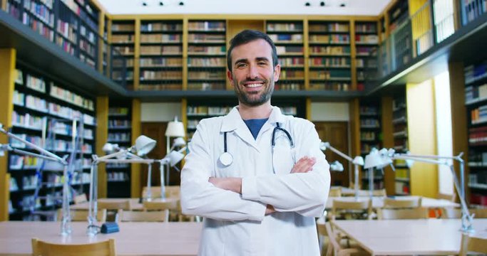 A young and beautiful doctor in a library smiling happy and holding books after doing a search and after studying. Concept: educational, portrait, library, and medical care and welfare