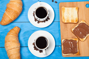 Two cups with coffee, croissant and toast with chocolate on a blue wooden table