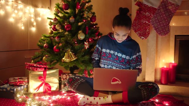 Young black woman is chatting online using laptop and sitting near decorated christmas tree