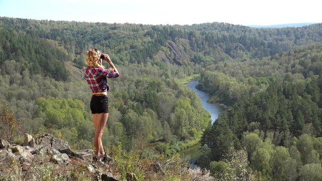 Young blonde woman tourist   on a cliff looking through binoculars on the landscape with the river