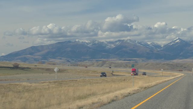 POV: Semi trucks and cars traveling along the busy countryside highway towards the magnificent snow-hooded Rocky Mountains. Wrinkled mountain range rising above the grassy prairie in Great Plains